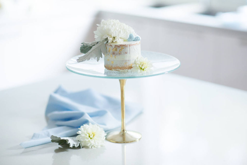 Baking Day Cake Stand resting upon hand cast bronze pedestals with small white cake topped with white and light blue flowers
