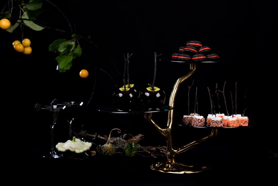 Finger food platters for Halloween. Dark and mysterious Halloween themed table setting consisting of deep night blue 3 tiered high tea stand on bronze tree, snack basket with chocolate, square plates with floral pattern with appetizers, fruit bowl with fruits inside and finger food platter in Filigree pattern. The composition is closed with a real tree branch on top.